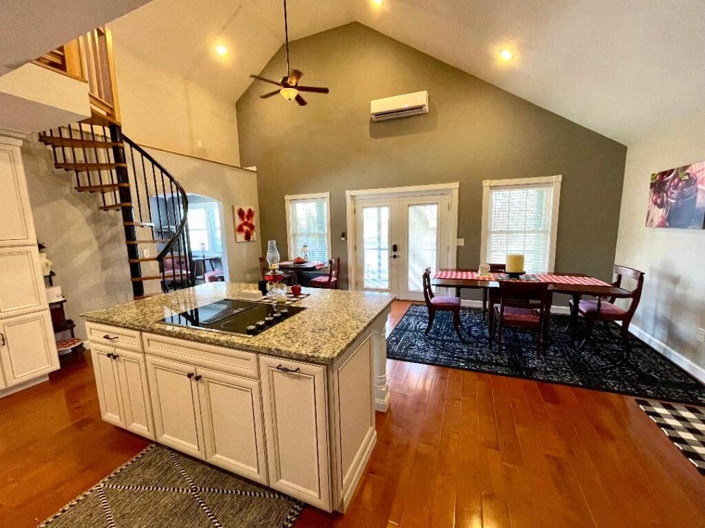 A spacious kitchen with a central island, leading to a dining area with a spiral staircase, in a room with vaulted ceilings and ample natural light.