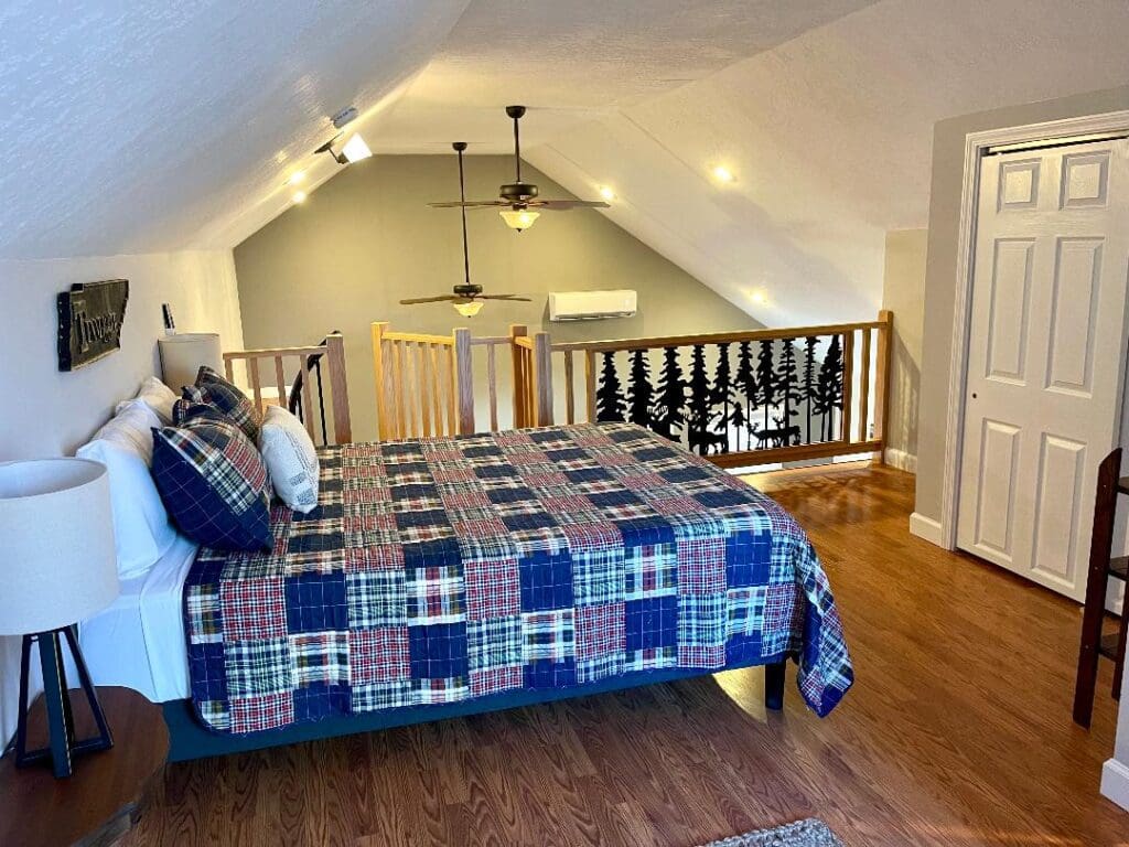 A cozy attic bedroom with a plaid bedspread, sloped ceilings, and a wooden railing overlooking the lower level.