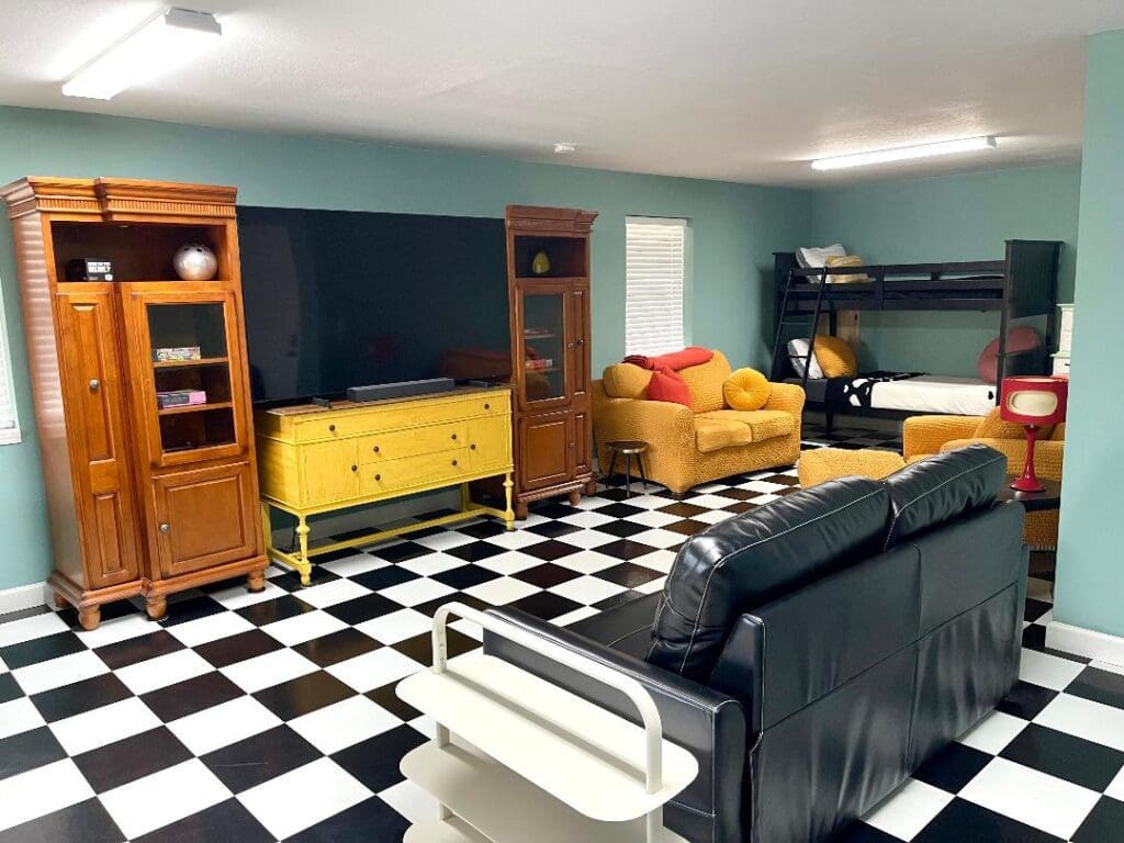 A colorful and eclectic living space with checkered flooring, a black leather sofa, wooden furniture, a bunk bed, and a large flat-screen tv.