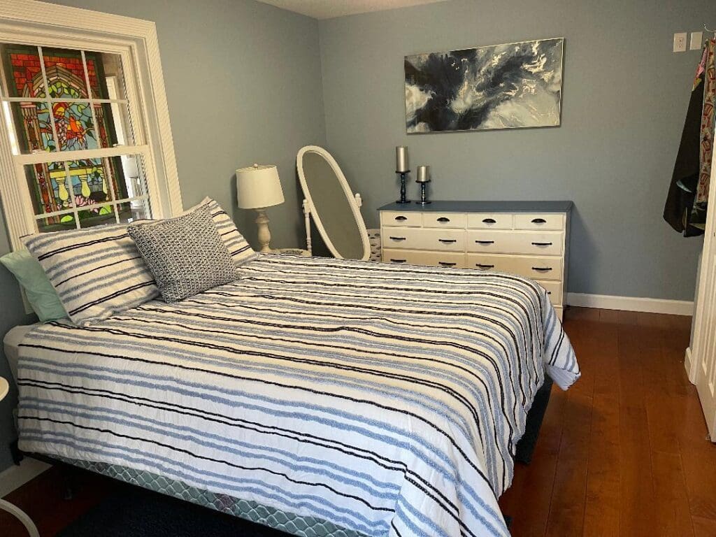 A neatly arranged bedroom with striped bedding, a white dresser, a wall painting, and a stained glass window.