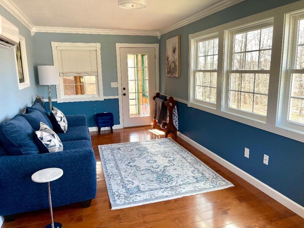 A cozy living room with blue walls, a blue sofa, and hardwood floors illuminated by natural light from surrounding windows.