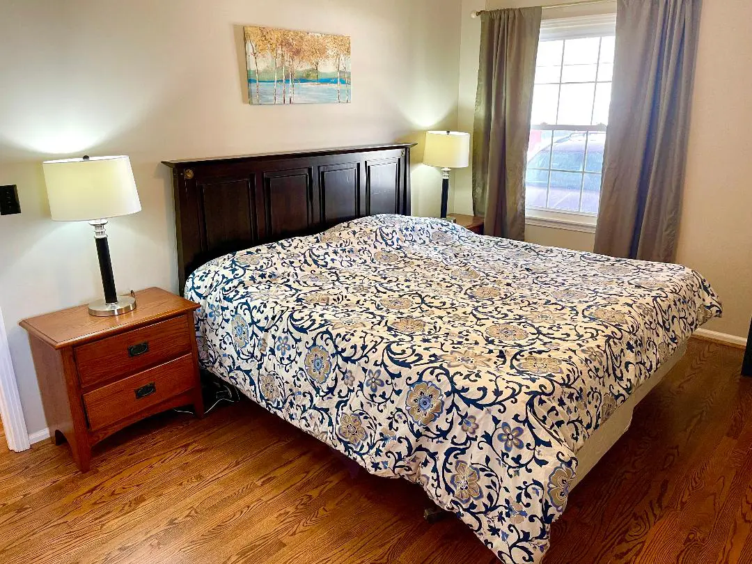 A well-lit bedroom with a queen-sized bed covered in a blue and white patterned bedspread, flanked by two lamps on matching wooden nightstands, and a wall art above the headboard.