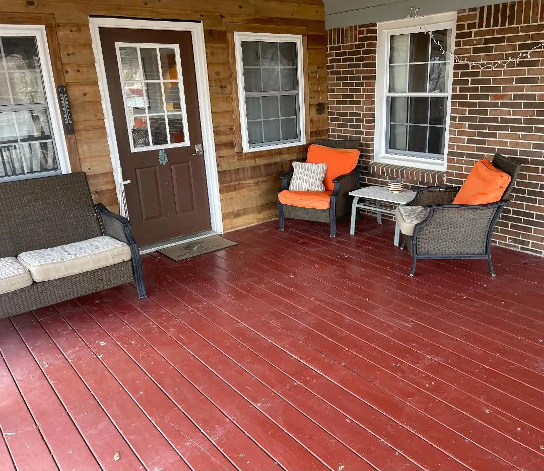 A cozy wooden porch with a red floor, furnished with a wicker sofa and two chairs with orange cushions.