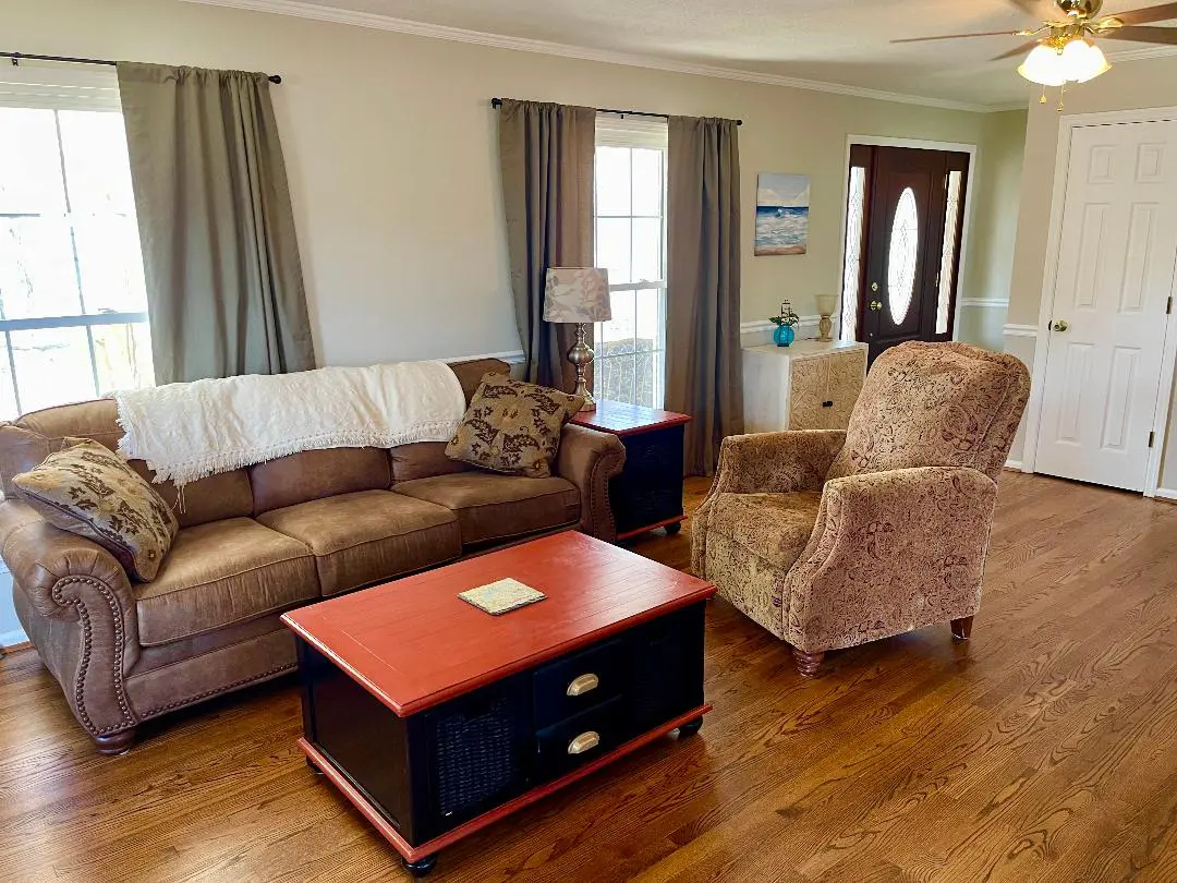 A well-lit living room featuring a brown leather sofa, patterned armchair, a red and black coffee table, and decorative elements, with hardwood flooring.