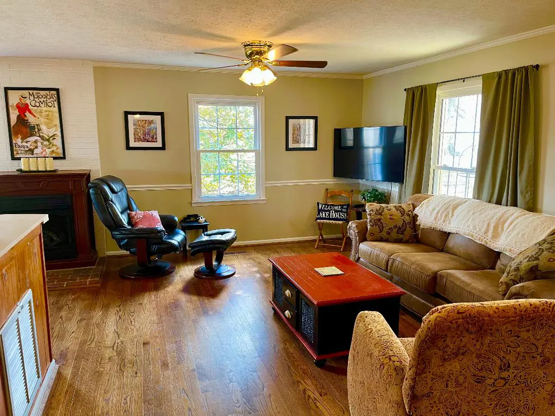 A cozy living room with a sofa, armchair, coffee table, and a mounted television, illuminated by natural light from two windows.