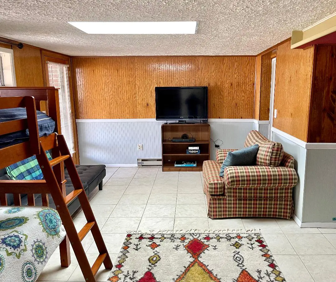 Interior of a modestly furnished room with a bunk bed, television, plaid chair, and patterned rug.