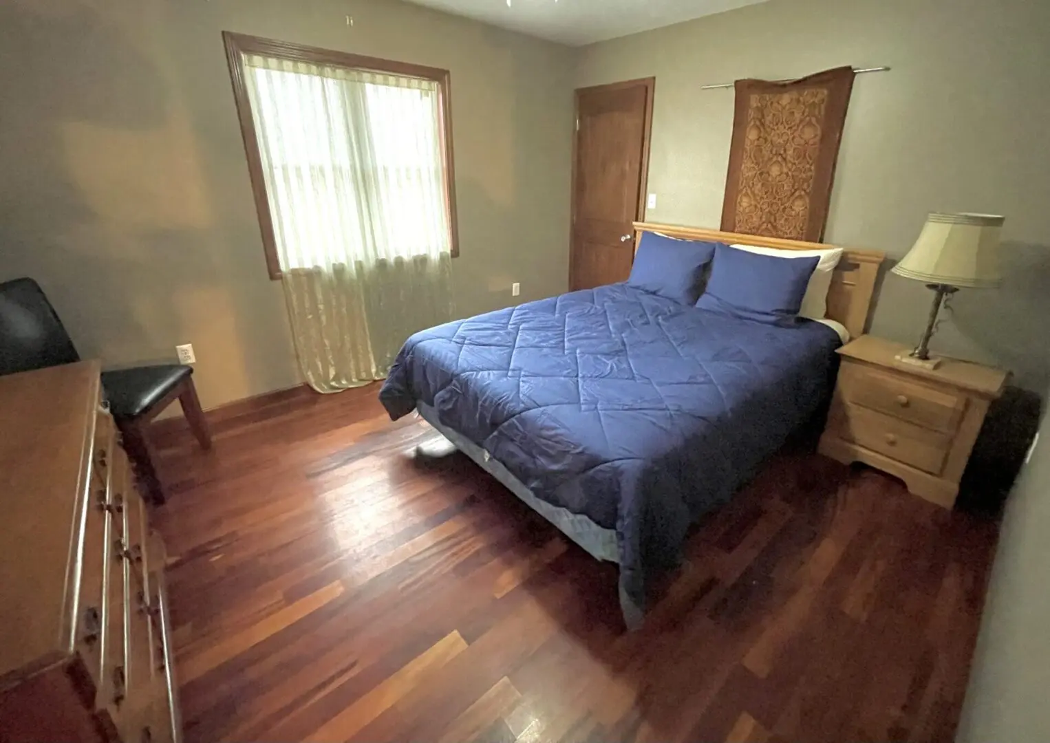 A tidy bedroom with a made bed, hardwood floors, and a window with sheer curtains.