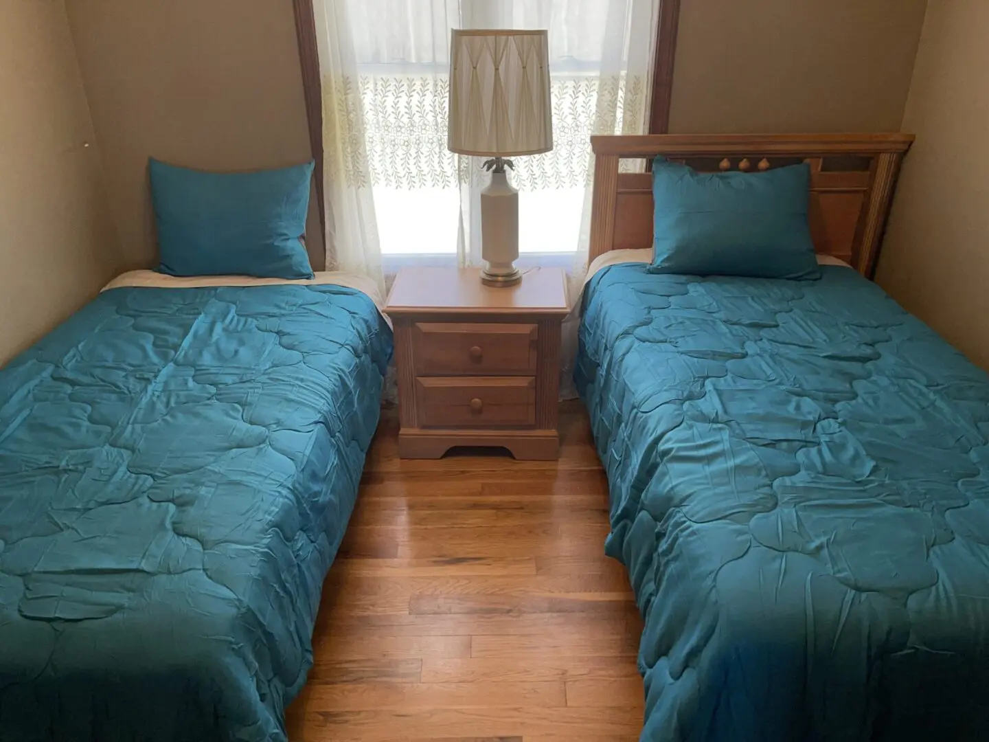 A simple bedroom with two twin beds covered in teal bedding, a nightstand in between, and a lamp on top.