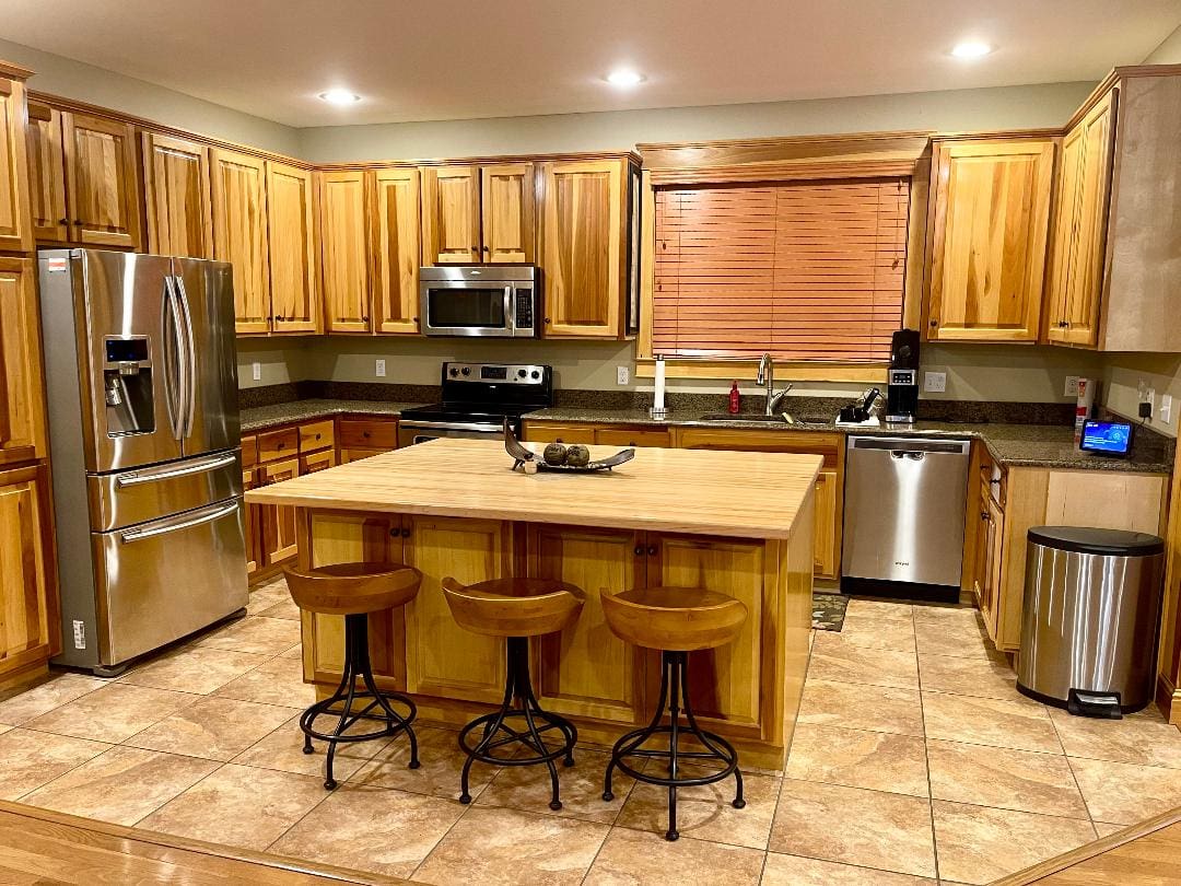 A modern kitchen with wooden cabinets, stainless steel appliances, a central island with bar stools, and tiled flooring.