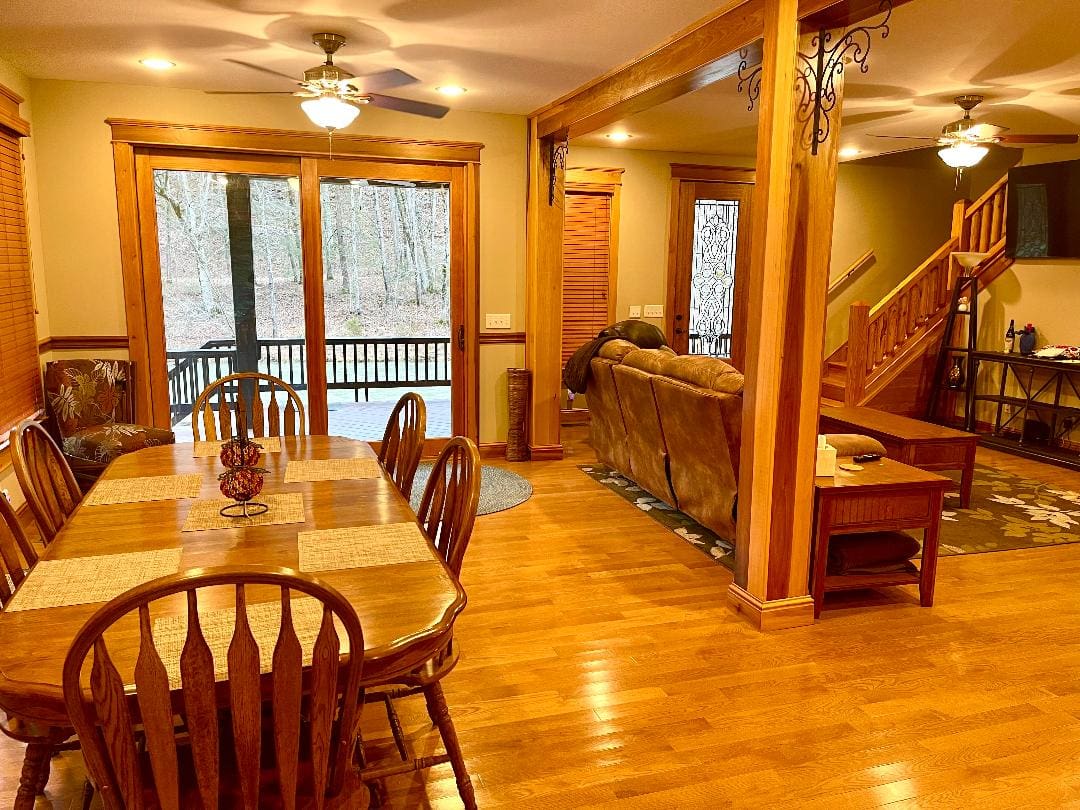 A cozy, well-lit dining area with a wooden table, chairs, and floors leading to a living space and a staircase, with views of the outdoors through large windows.