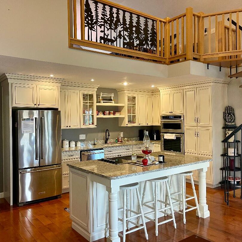 A modern kitchen with stainless steel appliances, white cabinetry, and a central island, under a balcony with decorative railing and a spiral staircase.